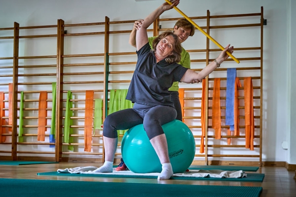 Eine Therapeutin zeigt einer Rehabilitandin die korrekte Haltung auf einem Pezziball